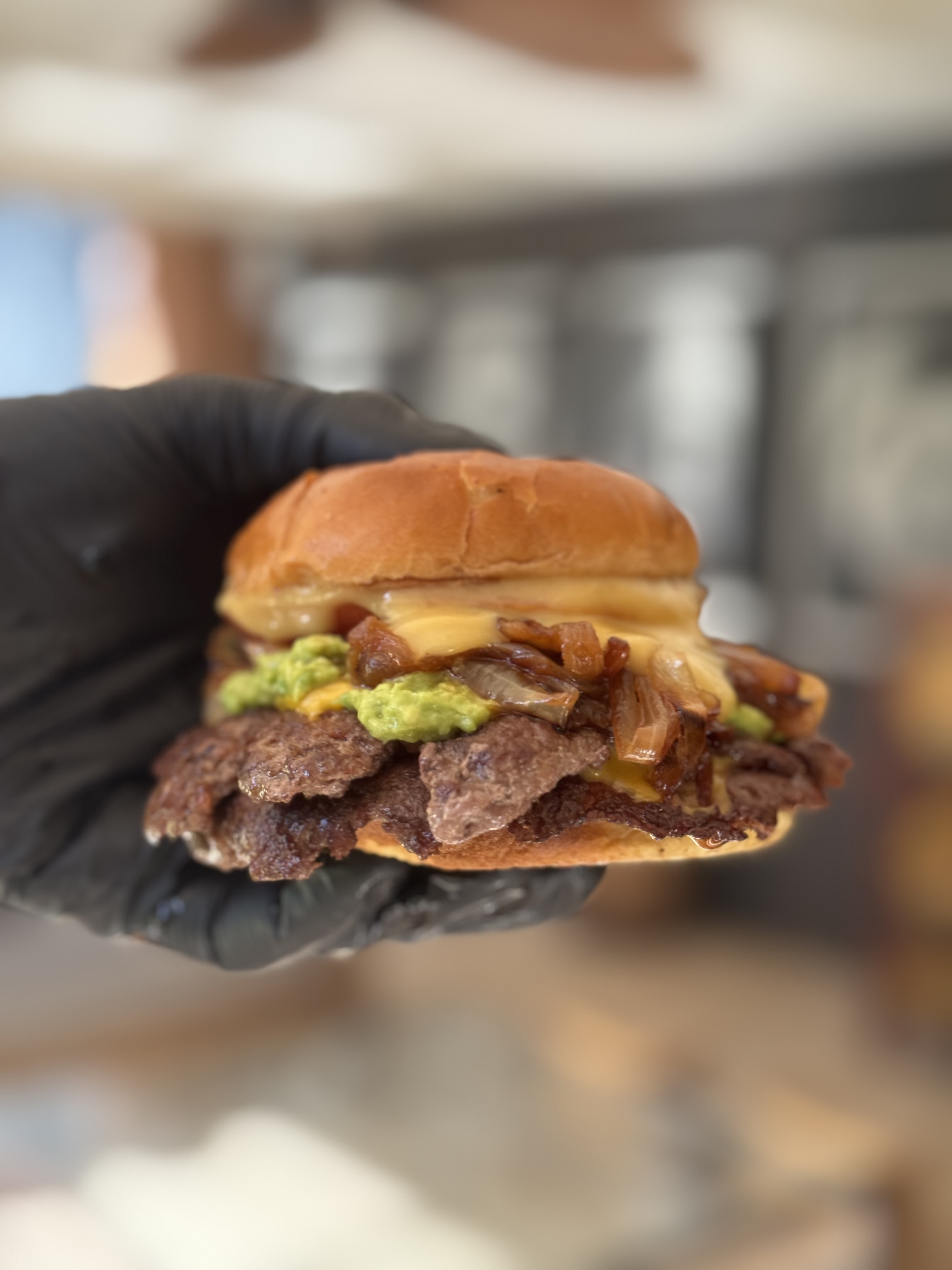 Hamburguesa smash doble con queso cheddar, cebolla caramelizada y palta, sostenida con guante negro
